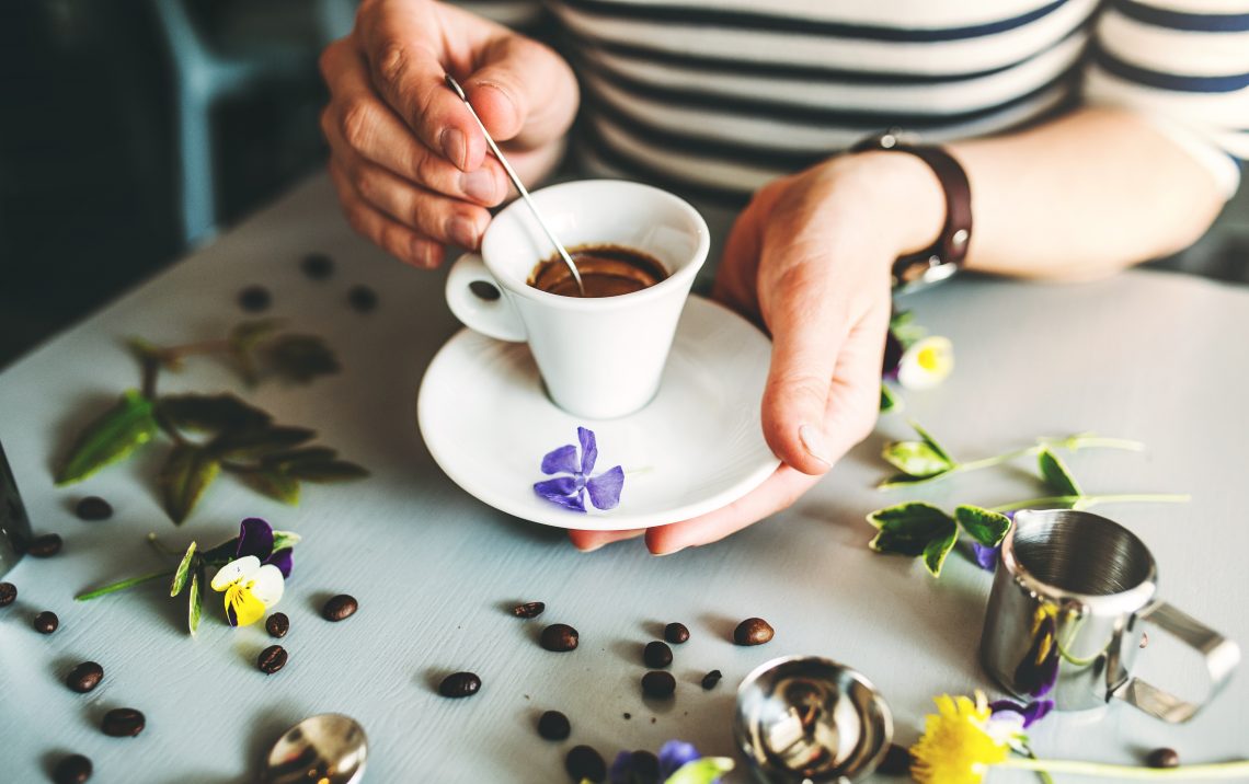 Kobieta trzyma w rękach filiżankę kawy. Na stole leżą płatki kwiatów i ziarna kawy
