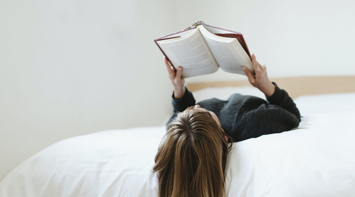 Tekst o pozytywnym wpływie czytania na zdrowie. Na zdjęciu: Kobieta leżąca na łóżku czytająca książkę - HelloZdrowie