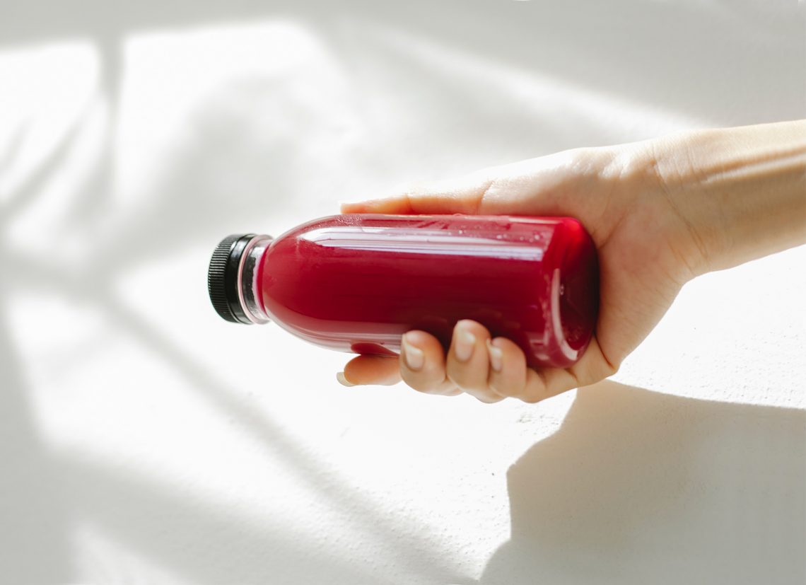 Tekst o wyborze najzdrowszego napoju owocowego w sklepie. Na zdjęciu: Dłoń trzymająca butelkę czerwonego płynu - HelloZdrowie