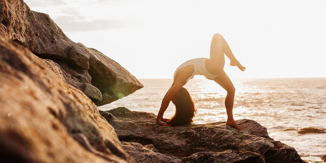 Tekst o treningu na plaży według Kasi Bigos. Na zdjęciu: Kobieta wykonująca jogę na skałach - HelloZdrowie