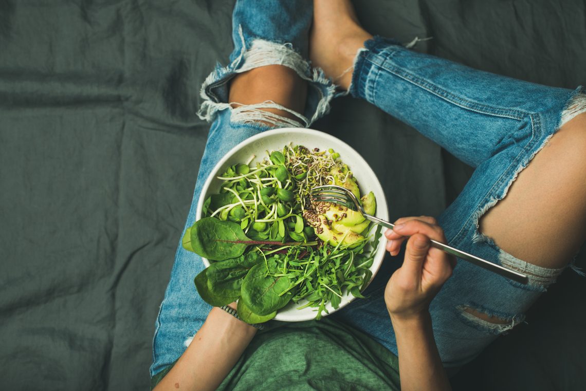 Tekst o zdrowych połączeniach żywieniowych w kuchni. Na zdjęciu: Osoba siedząca na podłodze jedząca miskę zielonych warzyw - HelloZdrowie