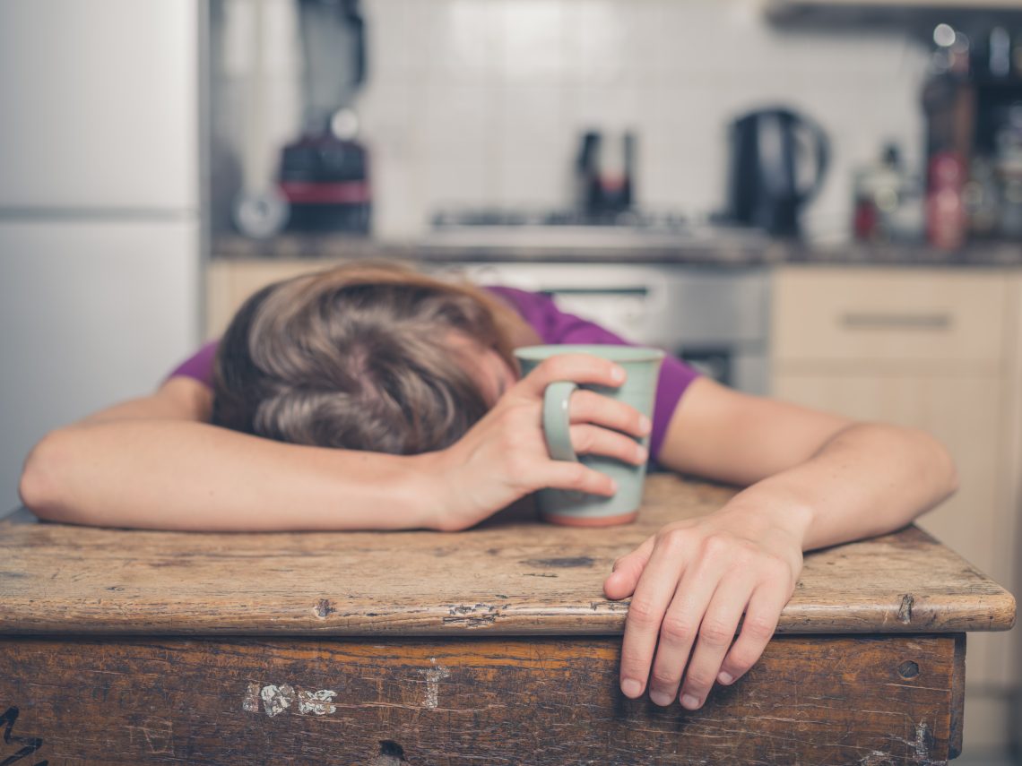Tekst o produktach, które zmniejszają zmęczenie. Na zdjęciu: Kobieta śpiąca na stole z głową na kubku - HelloZdrowie