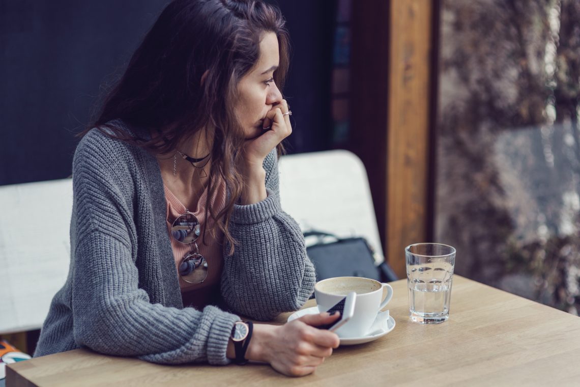 Tekst o zespole złamanego serca i jego przyczynach. Na zdjęciu: Kobieta siedząca przy stole z filiżanką kawy i telefonem - HelloZdrowie