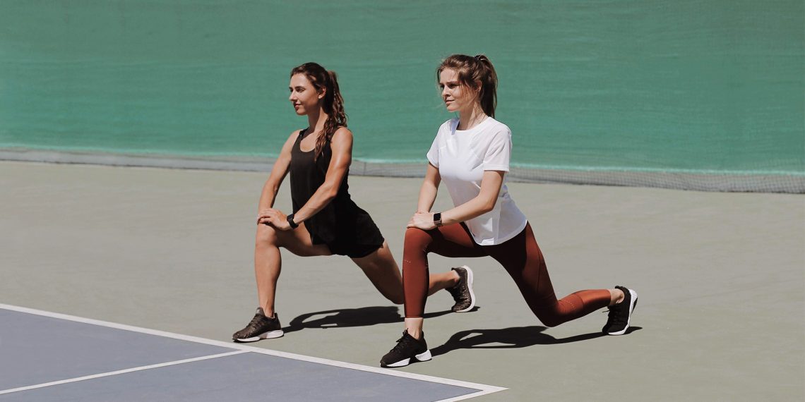Tekst o ćwiczeniach rozgrzewkowych przed bieganiem. Na zdjęciu: Dwie kobiety rozciągają się na kort tenisowy - HelloZdrowie