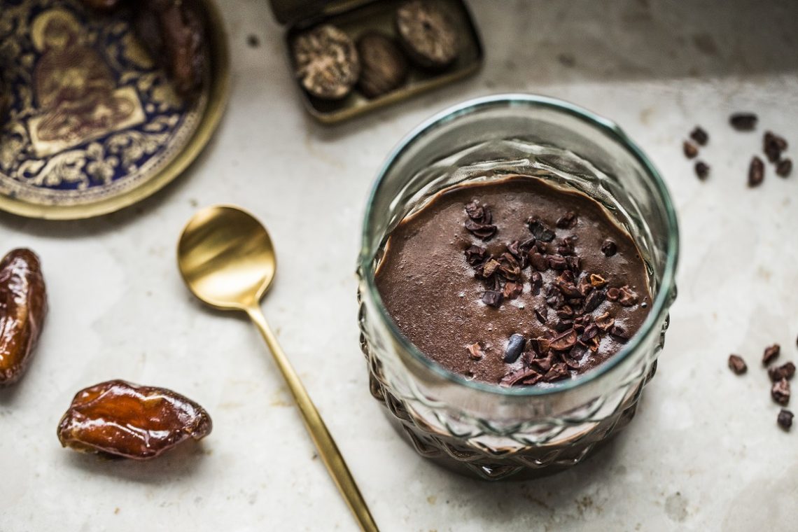 Tekst o przygotowaniu wegańskiej gorącej czekolady. Na zdjęciu: Szklanka puddingiem czekoladowym z łyżką oraz miseczka daktyli - HelloZdrowie