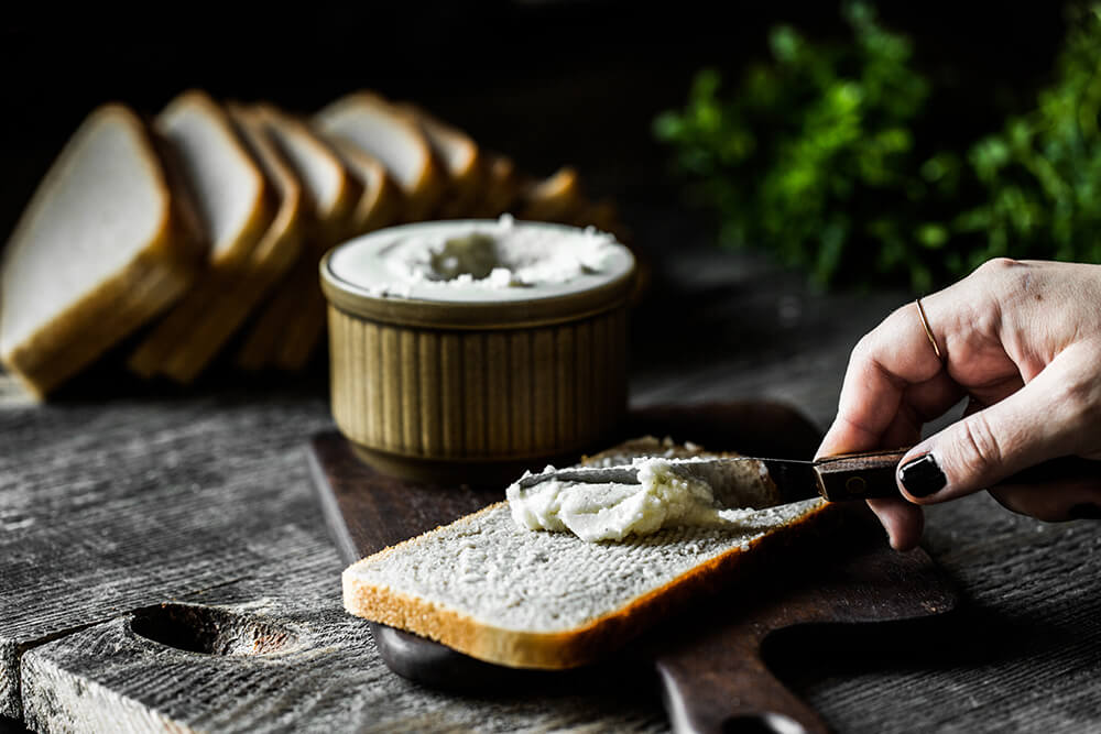 Tekst o domowym wegańskim maśle z kaszy jaglanej. Na zdjęciu: Osoba smarująca masło na kawałku chleba - HelloZdrowie