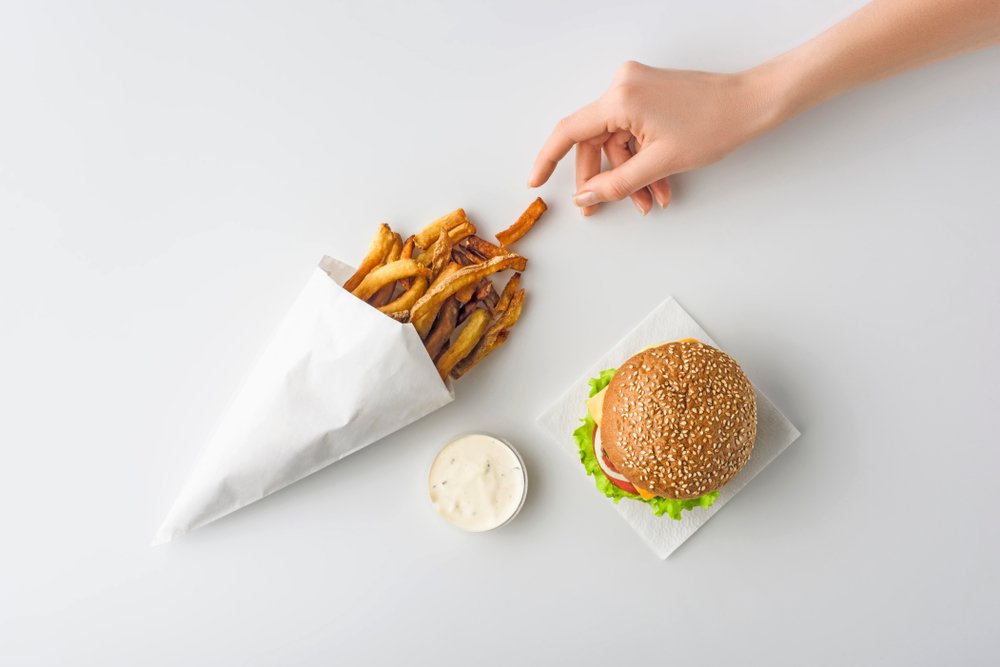 Tekst o wpływie tłuszczu na senność. Na zdjęciu: Ręka sięga po frytki w papierowym rożku obok burgera - HelloZdrowie