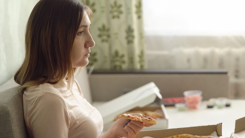 Smutna kobieta siedzi na kanapie z pizza i ogląda telewizję.