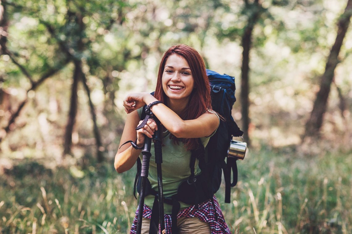 Tekst o korzyściach nordic walking nie tylko dla seniorów. Na zdjęciu: Kobieta z plecakiem i kijkami w lesie - HelloZdrowie