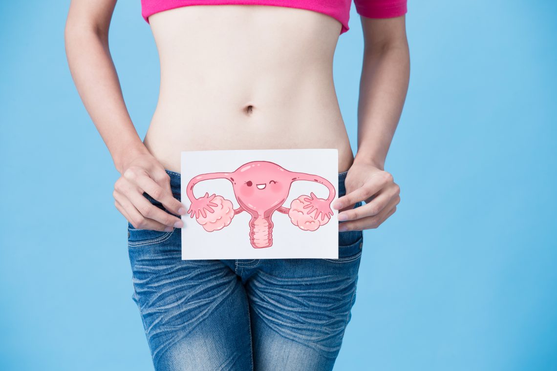 kobieta w jeansach i rózowej bluzce, z odkrytym brzuchem, trzyma kartkę z narysowanym układem rozrodczym (jajniki, szyjka macicy)