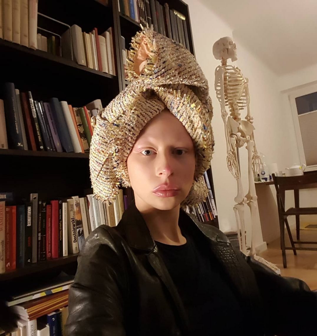 Zuzanna Bartoszek siedzi, na głowie ma zawiązaną tkaninę słomkową jak zwykłą chustę. Za nią regał z książkami oraz szkielet medyczny człowieka
