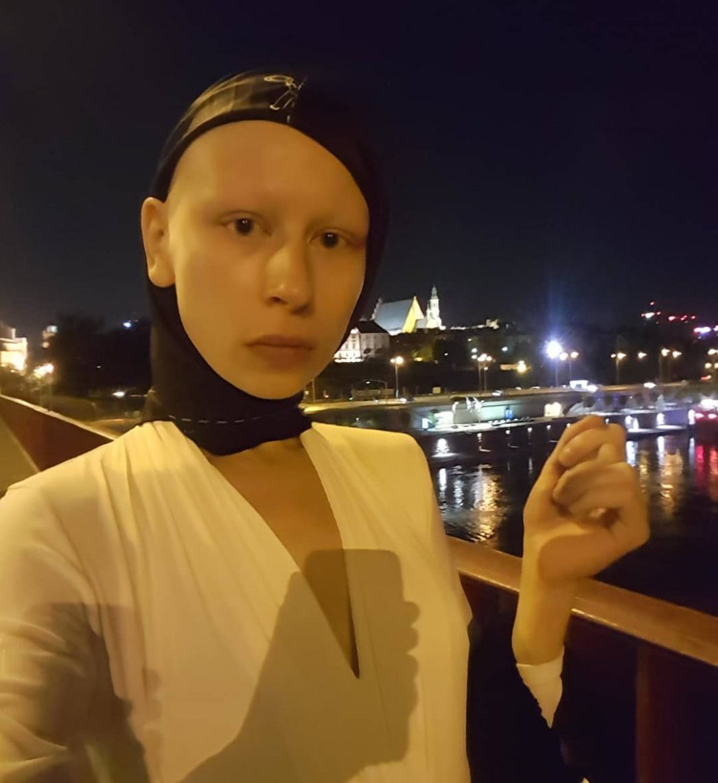 Zuzanna Bartoszek stoi nocą na moście, w tle lampy miasta. Ubrana na biało, na głowie ma czarną kominiarkę z wycięcięm na całą twarz
