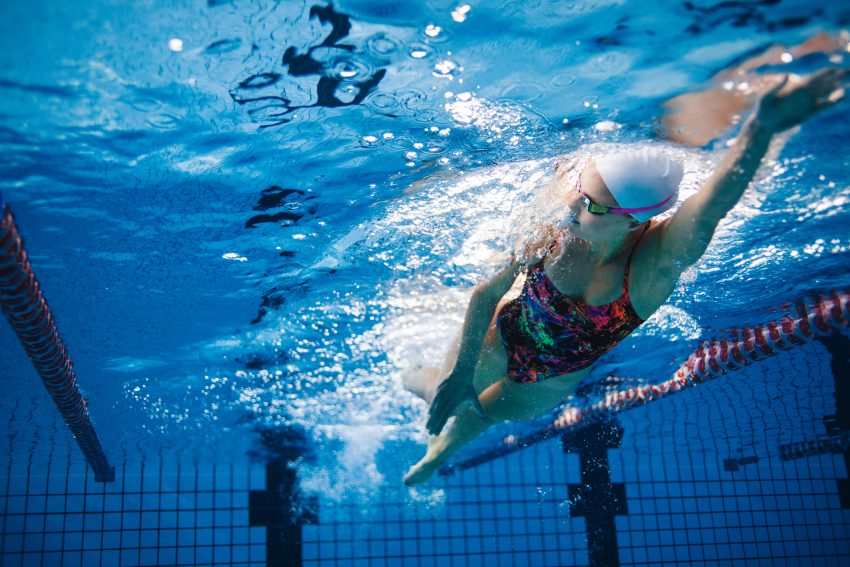 kobieta płynie kraulem na sportwym basenie, ubrana w kolorowy, jednoczęśowy kostium, czepek oraz okulary do pływania. Zdjecie zrobione spod wody.