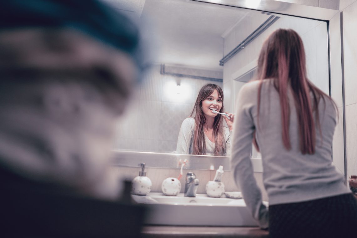 Tekst o wpływie higieny jamy ustnej na nadciśnienie. Na zdjęciu: Kobieta myjąca zęby przed lustrem - HelloZdrowie