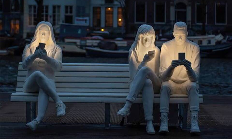 Białe rzeźby przedstawiające troje młodych lduzi siedzących na ławce i patrzących w telefony. Telefony oświetlają ich twarze.