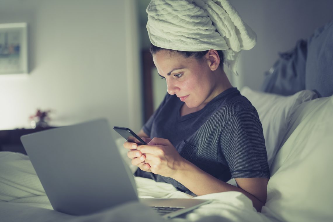 Kobieta leży na łóżku przed laptopem, ma na głowie ręcznik