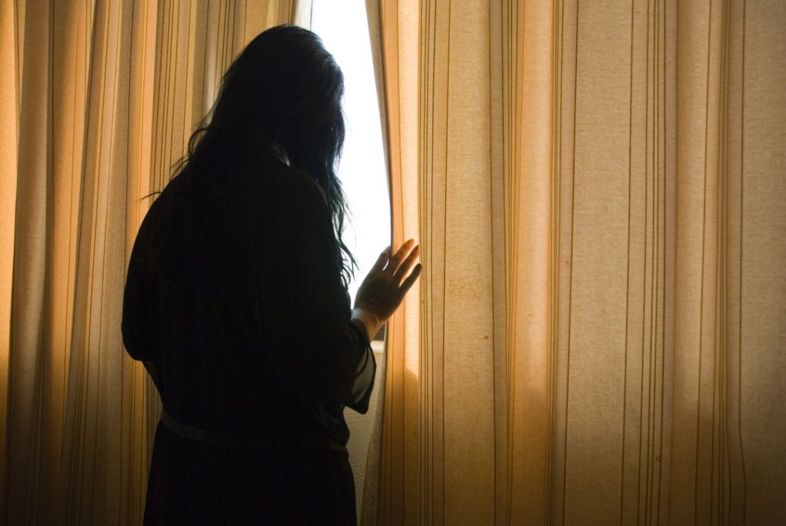 Kobieta cierpiąca na agorafobię wyglądająca przez okno zza firanki