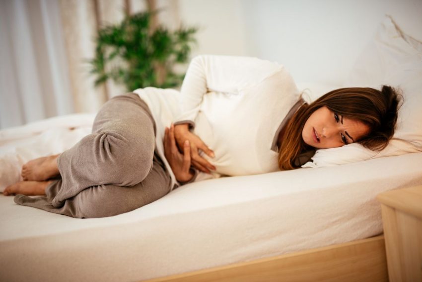 Kobieta w białej bluzce leży na lóżku w ubraniu i trzyma się za brzuch. Ma wykrzywioną twarz