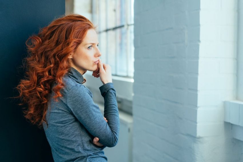 Niezadowolona kobieta w rudych kręconych włosach stoi w przedpokoju i patrzy przez okno.