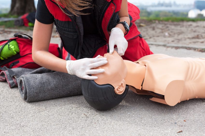 Na zdjęciu: Osoba w czerwonym kamizelce i rękawiczkach ćwiczy resuscytację krążeniowo-oddechową na manekinie - HelloZdrowie