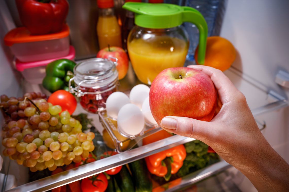 Tekst o prawidłowym przechowywaniu różnych produktów spożywczych. Na zdjęciu: Ręka trzymająca jabłko przed lodówką pełną jedzenia - HelloZdrowie