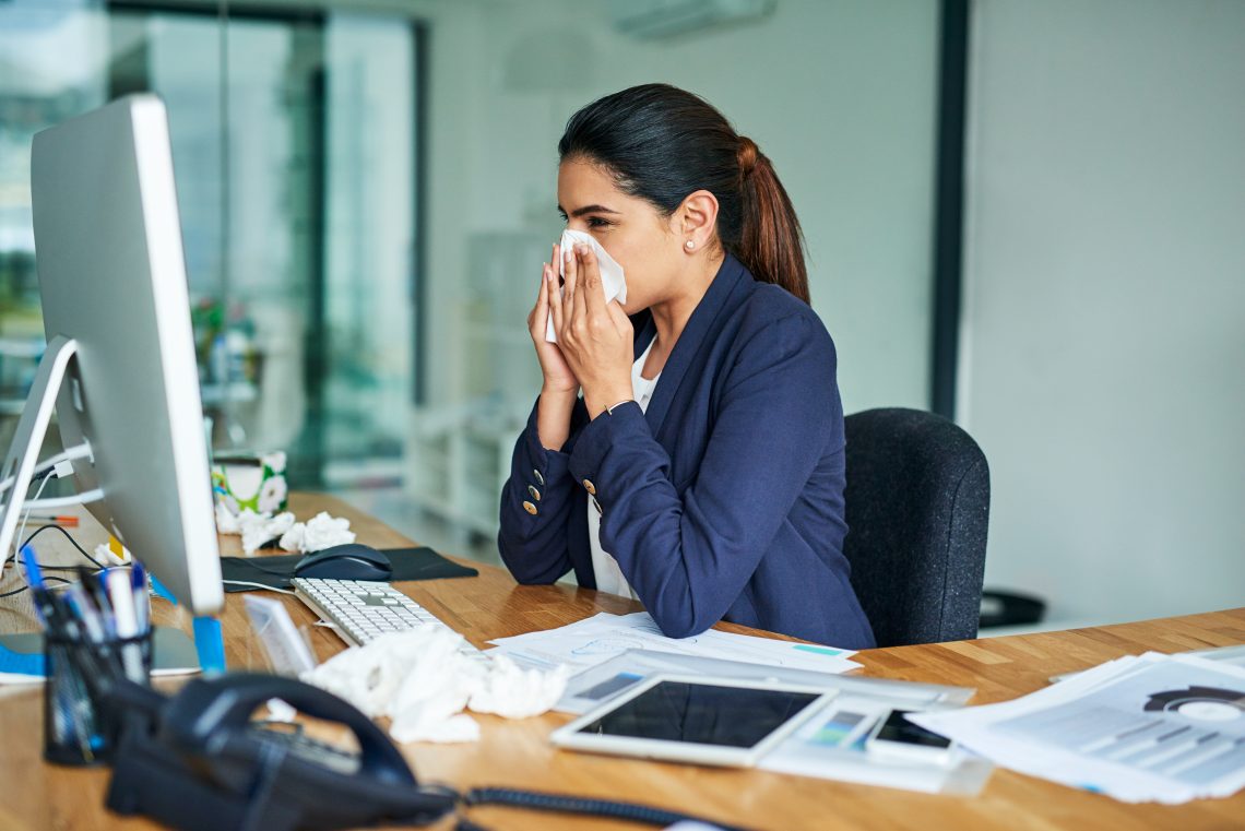 kobieta siedzi w pracy przy biurku przed komputerem i wydmuchuje nos w chusteczkę