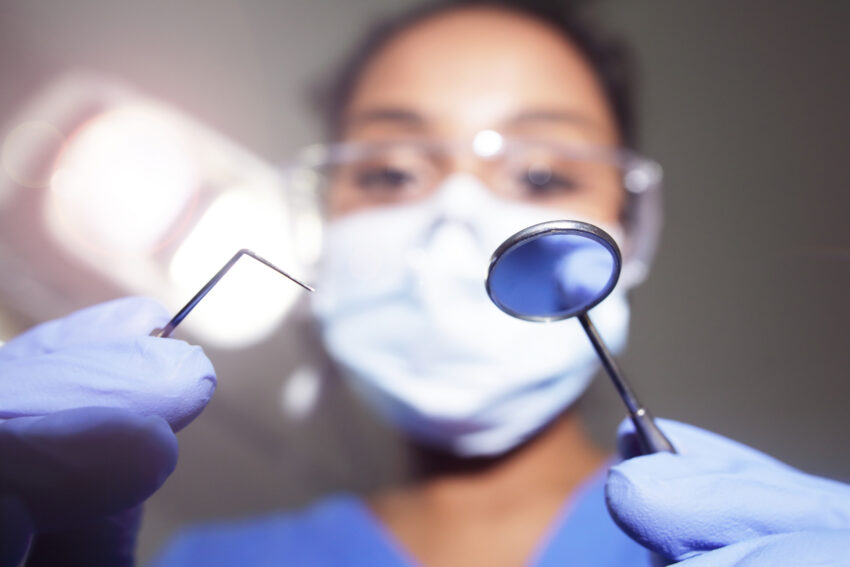 Na zdjęciu: KObieta stomatolog pochyla się nad pacjentem, na pierwszy plan wysuwają się narzędzia stomatologiczne