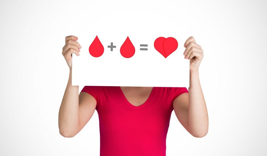 Kobieta stoi z kartką, na której narysowane są krople krwi, które po zsumowaniu dają serce