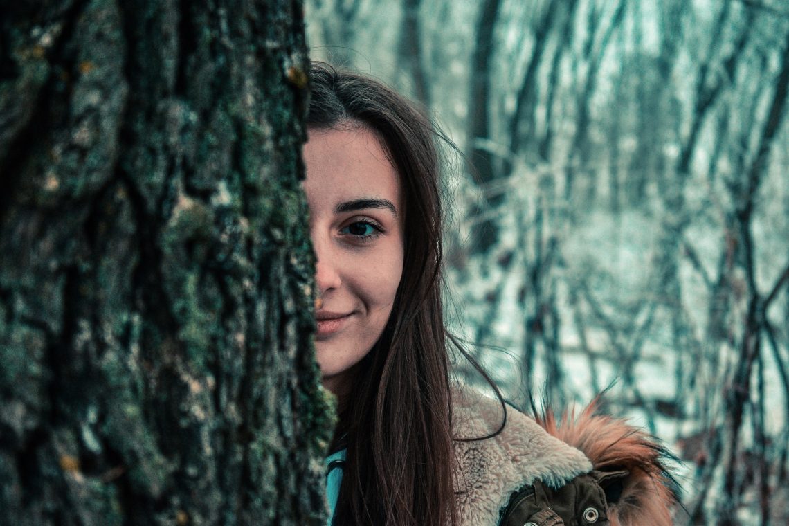 Tekst o domowych sposobach na suche usta. Na zdjęciu: Kobieta uśmiechająca się zza drzewa - HelloZdrowie