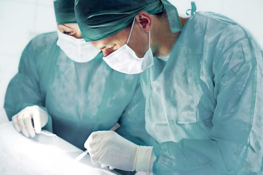 Dwóch chirurgów za pomocą lanceta wykonuje zabieg. Mają na sobie zielone fartuchy.