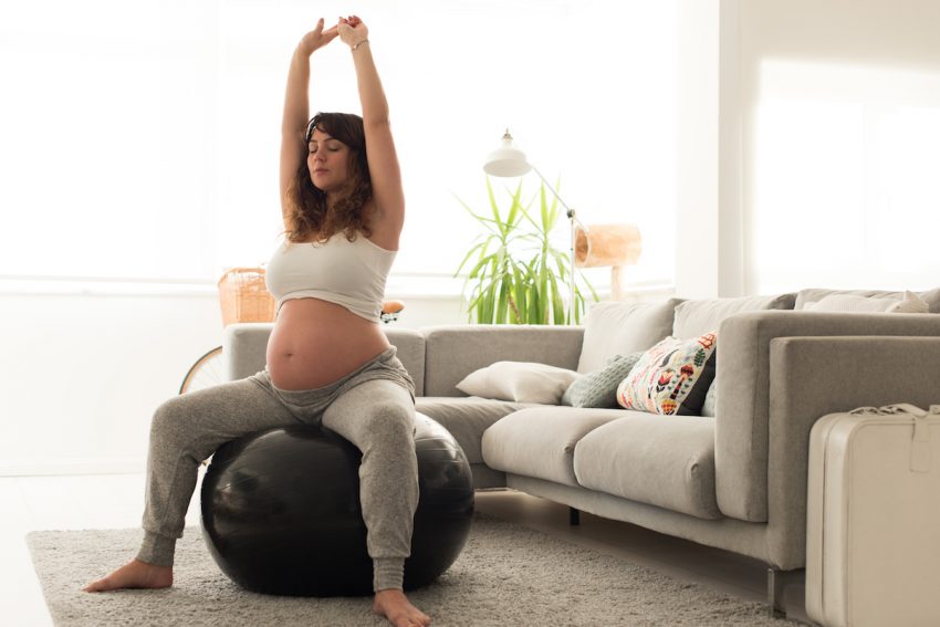 Młoda kobieta w ciąży siedzi na piłce do ćwiczeń i wyciąga się w górę.