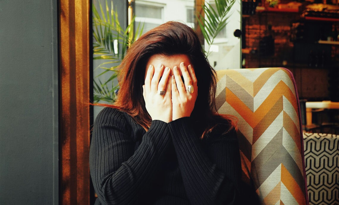 Afonia – czy można stracić głos z powodu silnego stresu?; na zdjęciu kobieta zasłaniająca dłońmi twarz- Hello Zdrowie