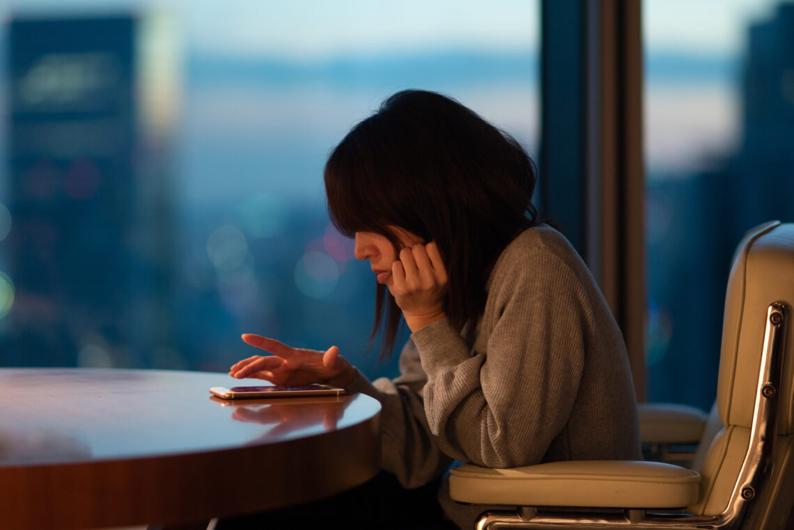 Na zdjęciu: Kobieta siedzi zgarbiona przy stole, używa telefonu komórkowego