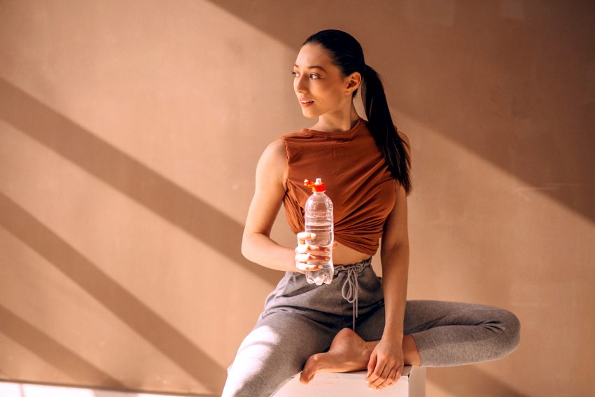 Tekst o ćwiczeniach mięśni dna miednicy i nietrzymaniu moczu. Na zdjęciu: Kobieta siedząca na sześcianie trzymająca butelkę wody - HelloZdrowie