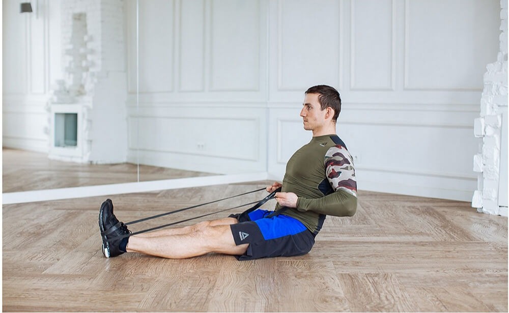 Tekst o zastosowaniu gum w treningu siłowym. Na zdjęciu: Mężczyzna siedzący na podłodze z opaską oporową - HelloZdrowie