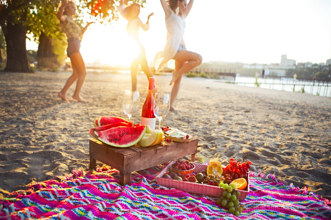 Trzy kobiey tańczą na plaży. Przed nimi koc a na nim owoce i drink