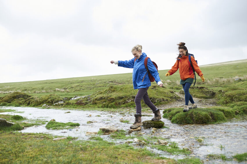 Na zdjęciu dwie kobiety ubrane w kurtki i buty trekkingowe uśmiechają się, przechodząc przez rzekę po kamieniach; otaczają je pokryte trawą wzgórza.