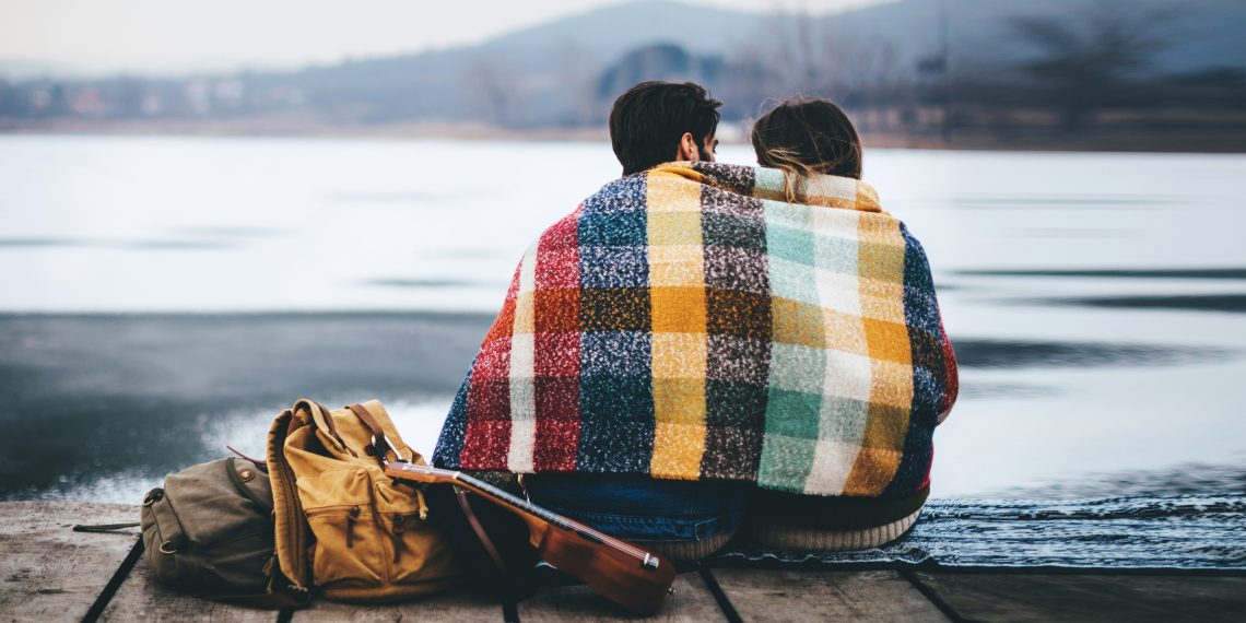 Tekst o różnicach w odczuwaniu temperatury przez płcie. Na zdjęciu: Mężczyzna i kobieta siedzący na pomost z gitarą i plecakiem - HelloZdrowie