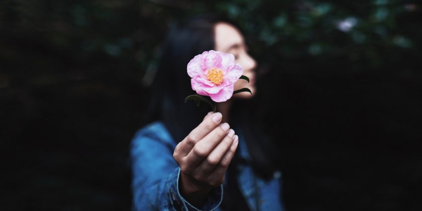 Kobieta patrzy w bok, wyciąga rękę, w której trzyma kwiatek