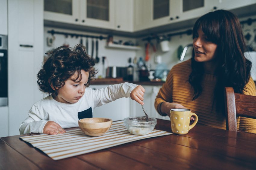 Kobieta z dzieckiem siedzą przy stole w kuchni. Mama pije kawę a dziecko je z dwóch salaterek śniadanie