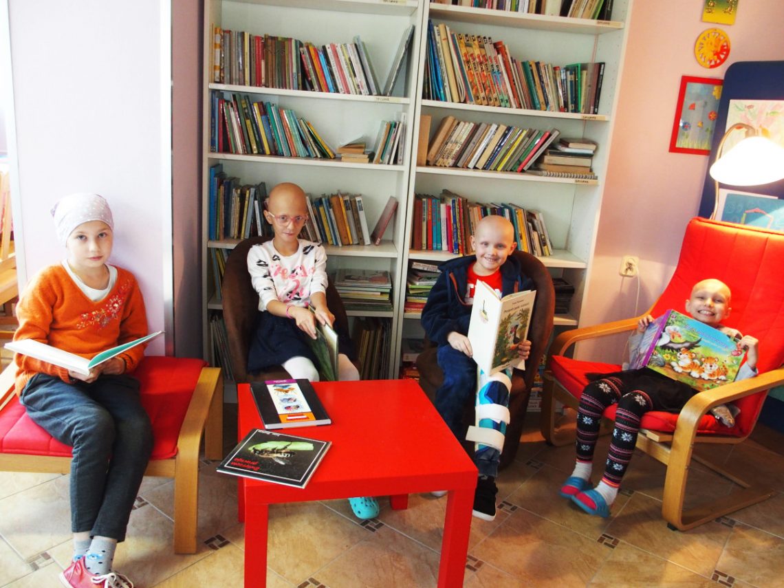 Dzieci z nowotworem siedzą przy stoliku na krzesełkach i czytają książki, uśmiechają się