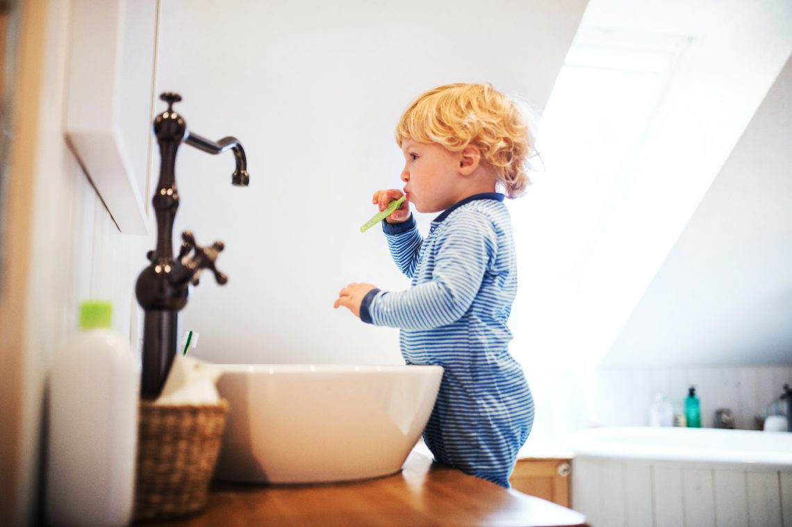 Mały chłopiec stoi na krzesełku przy umywalce, patrzy w lustro i myje zęby