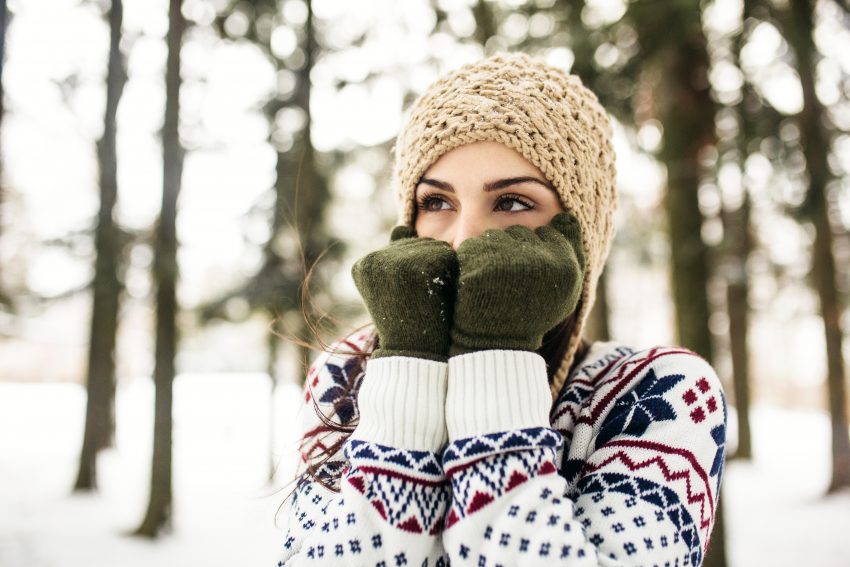 Kobieta stoi w lesie, jest zima i śnieg. Ubrana jest w sweter, czapkę i rękawiczki. Ręce trzyma przy tywarzy, patrzy się w bok