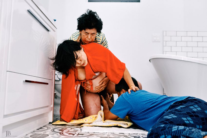 Tekst o konkursie fotografii porodowej i nagrodzonych zdjęciach. Na zdjęciu: Kobieta klęcząca na podłodze z mężczyzną i kobietą z tyłu - HelloZdrowie