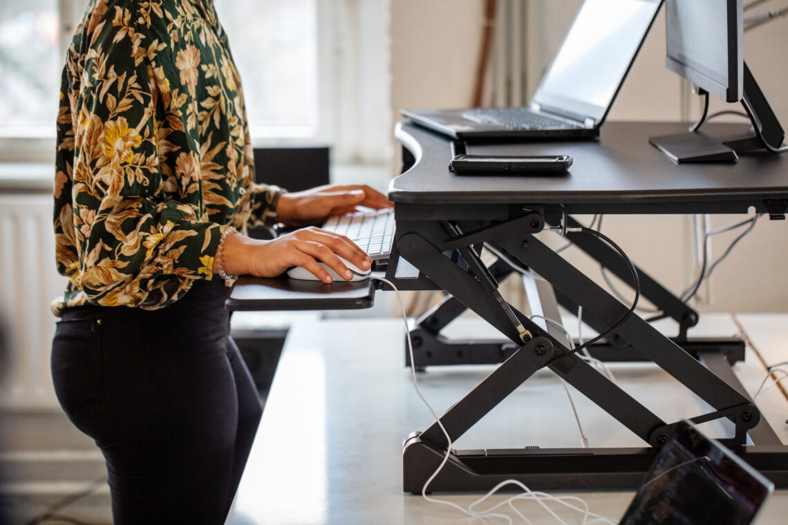 Kobieta stoi przy biurku i pracuje przy laptopie; ergonomia pracy polega tu na dostosowaniu wysokości komputera, klawiatury i myszki.