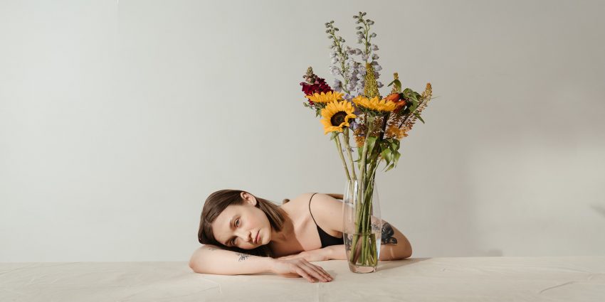 Kobieta siedzy na krześle, jednocześńie leżąc na stole. Obok niej stoi wazon kwiatów