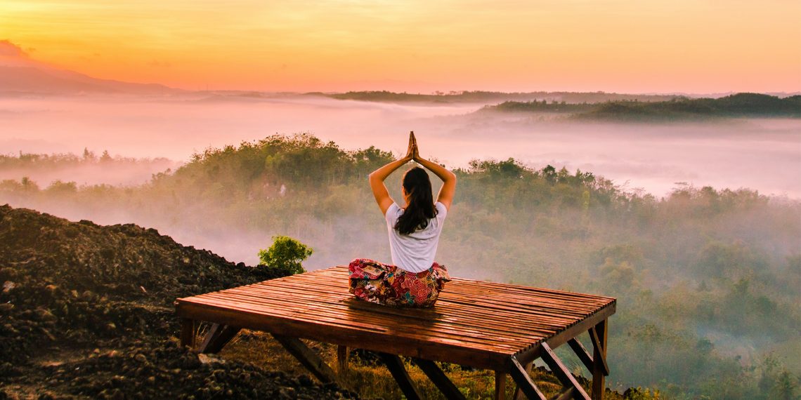 Tekst o korzyściach z medytacji i jak zaczynać. Na zdjęciu: Kobieta siedząca na drewnianej platformie z złożonymi rękoma - HelloZdrowie