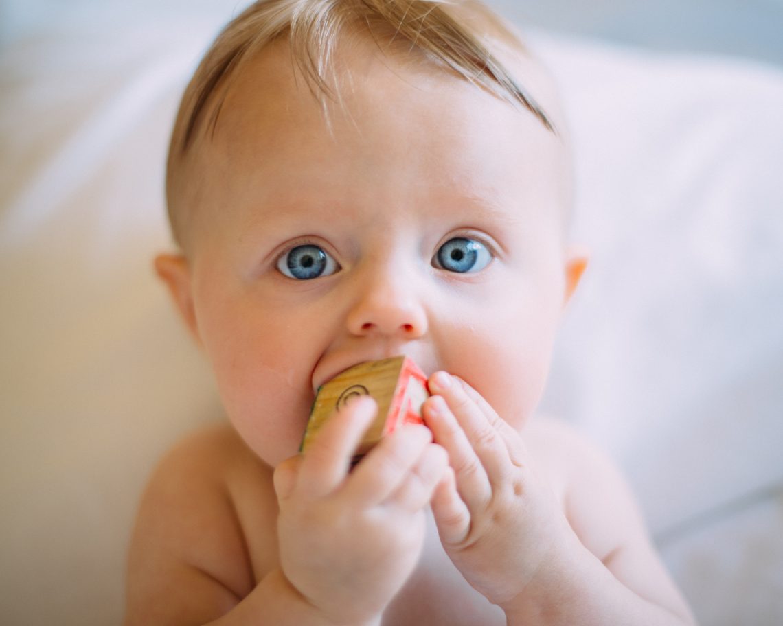 Tekst o rozszerzaniu diety niemowląt i zaleceniach. Na zdjęciu: Dziecko o niebieskich oczach gryzące klocek - HelloZdrowie