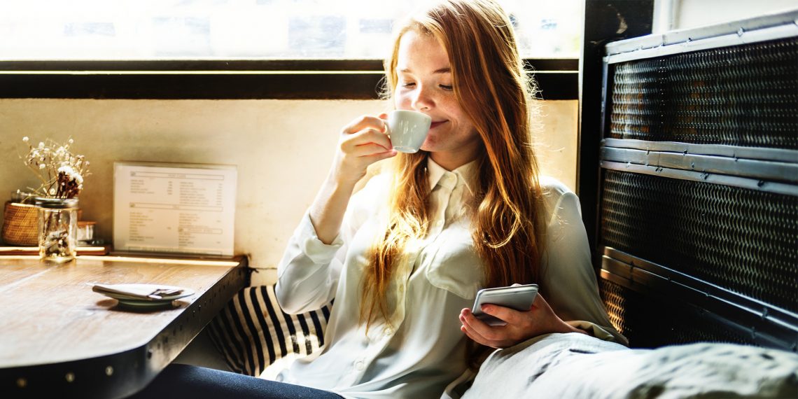 Tekst o zdrowotnych korzyściach płynących z picia kawy. Na zdjęciu: Kobieta pijąca kawę i korzystająca z telefonu - HelloZdrowie