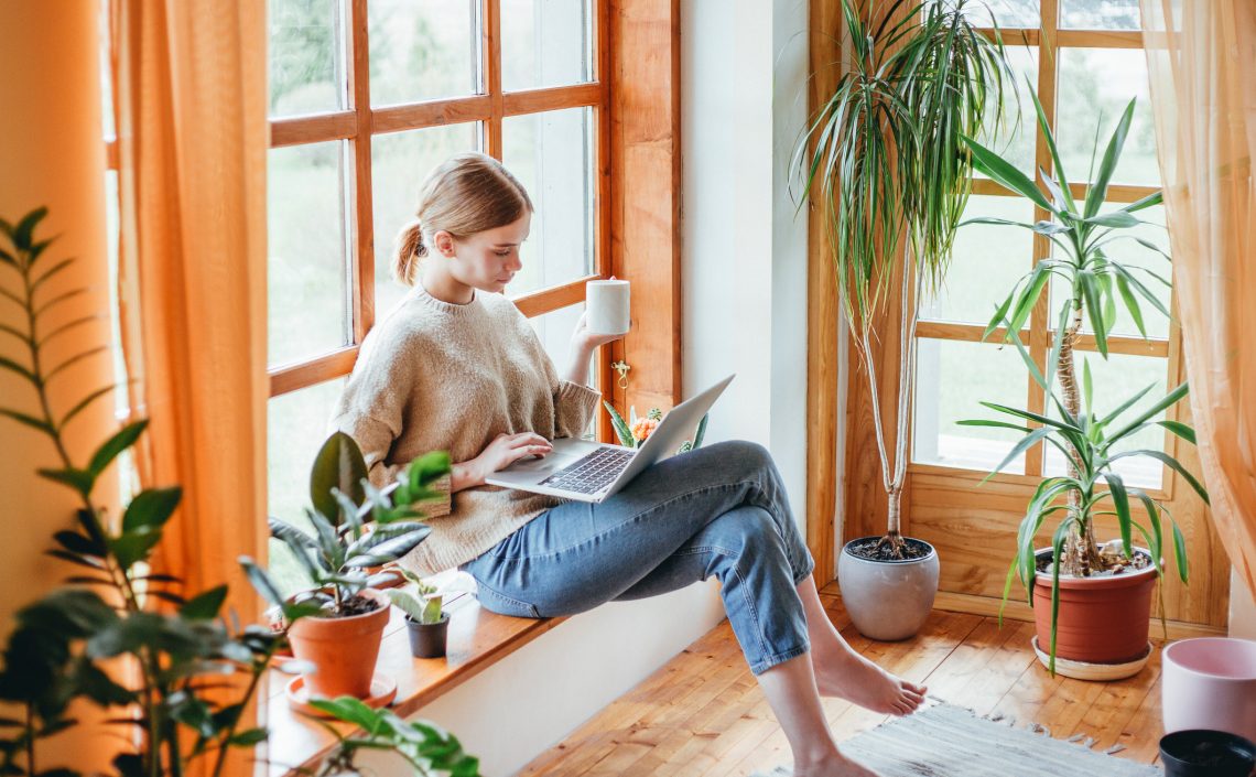 Tekst o zachowaniu wagi podczas siedzenia w domu. Na zdjęciu: Kobieta siedząca na parapetem z laptopem - HelloZdrowie